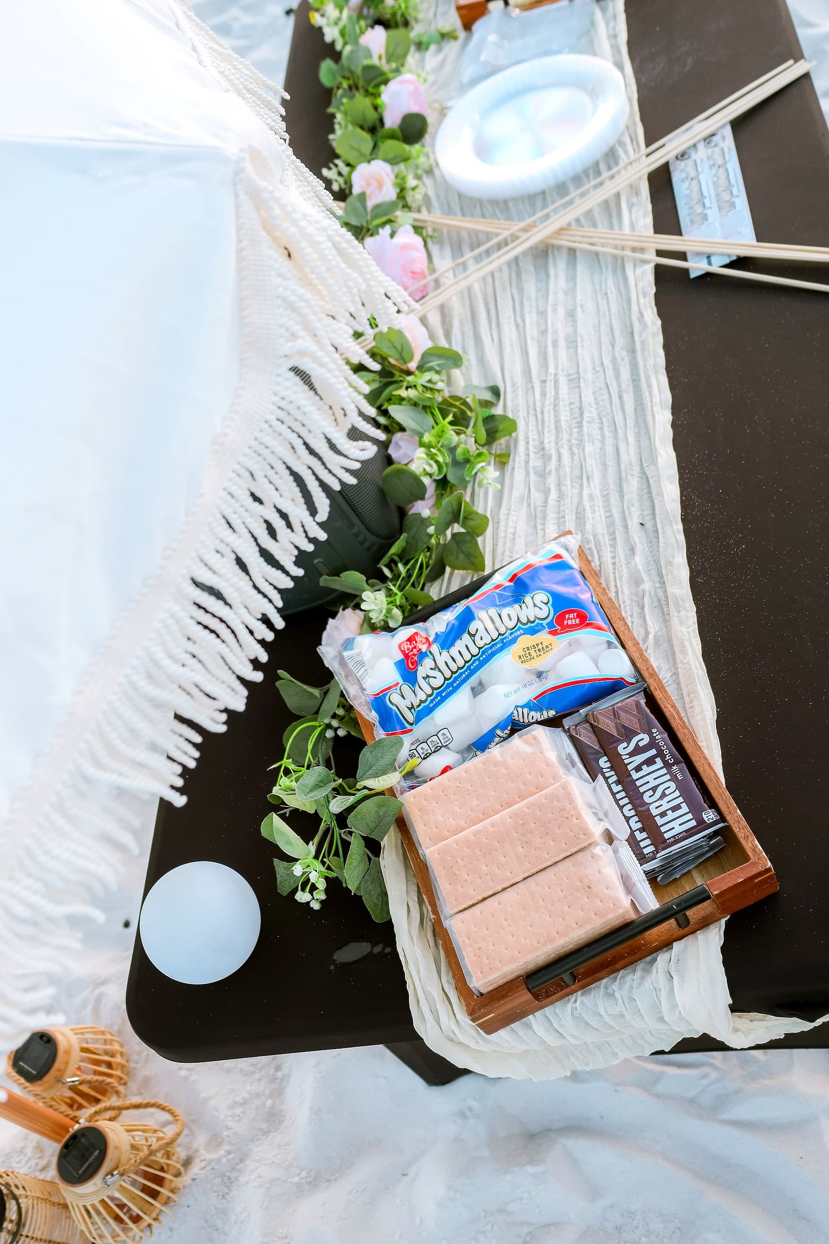 A styled beach bonfire table with fresh florals, a gourmet s'mores spread, greenery garland and a speaker on the white sand